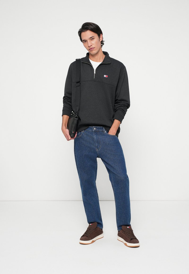 Junger Mann, der einen dunkelgrauen Pullover mit Viertelreißverschluss, blaue Jeans, braune Sneakers trägt und eine schwarze Umhängetasche hält, steht vor einem weißen Hintergrund.