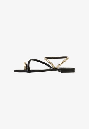 Flad sort sandal med minimalistisk gyldent tå-loop og asymmetrisk ankelrem, set fra siden på hvid baggrund.