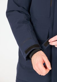 Marinblå vattentät jacka med kardborremudd, som har flera fickor och "W-PRO 10000"-logotyp på ärmen. Texturerat tyg.