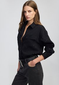 Czarna koszula z teksturowanym wzorem, z zapięciem na guziki z przodu i dwiema kieszeniami na piersi. Noszona z ciemnymi dżinsami i czarnym paskiem.