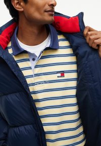 Man draagt een gestreept geel-blauw Tommy Jeans poloshirt onder een donkerblauwe jas met een rode binnenkraag.