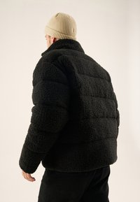 Svart, myk pufferjakke i fløyel, med teksturert materiale, høy krage og sydd paneler. Kombinert med en lys beige ribbestrikket lue.