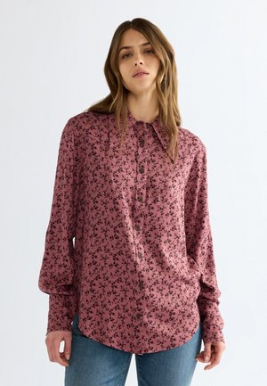 Junge Frau mit hellbraunen Haaren trägt ein langärmliges pinkes Shirt mit schwarzem Blumenmuster und blaue Jeans vor einem einfarbigen hellen Hintergrund.