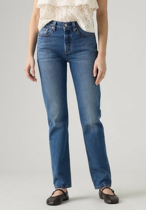 Kvinde iført lige blå jeans, cremefarvet blondebluse og sorte flade sko, stående mod en ensfarvet baggrund.
