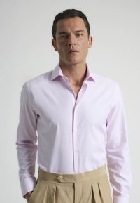 Camisa de manga larga rosa claro con cuello estructurado, sutil brillo y frente con botones. Combinada con pantalones beige plisados.
