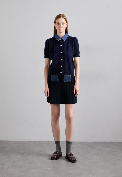 Navy cable-knit short-sleeve cardigan with denim collar, paired with a black pleated skirt featuring blue pockets, and dark loafers with studs.
