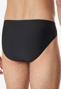 Slip da nuoto neri con sottili strisce bianche, realizzati in un tessuto morbido e con un taglio a vita bassa che offre una copertura minima sul retro.