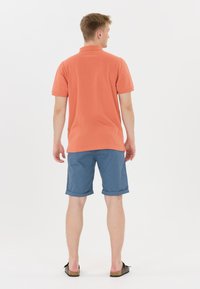 Korallfärgad pikétröja med korta ärmar, matchad med blå uppvikta shorts och svarta sandaler, sedd framifrån. Minimalistisk design.