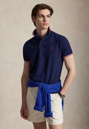 Fiatal férfi tengerészkék pólóingben piros logóval, bézs rövidnadrágban, kék pulóverrel a deréknál megkötve és karórával, oldalra néz.