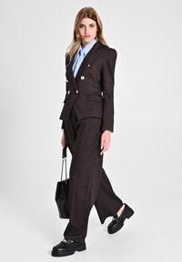 Blazer doppiopetto marrone, camicia azzurra chiara, pantaloni marroni larghi, mocassini neri spessi e una borsa a tracolla nera trapuntata.
