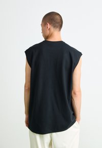 Moški s kratkimi lasmi, oblečen v črn brezrokavnik in bele hlače, stoji obrnjeno stran od preproste svetle ozadja.