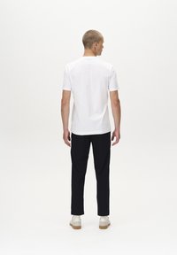 Camiseta blanca de manga corta con cuello redondo, combinada con pantalones negros y zapatillas blancas, vista desde atrás. Textura suave.