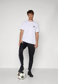 Camiseta blanca de manga corta con pequeño logo, combinada con pantalones deportivos negros con franjas blancas laterales, de pie sobre un balón de fútbol.