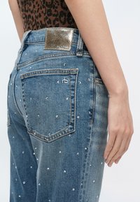 Femme portant un jean denim bleu avec de petites clous argentés et un patch en cuir doré à la taille, assorti à un haut à imprimé léopard.