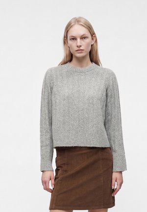 Jeune femme portant un pull en tricot gris et une jupe en velours côtelé marron, debout devant un fond clair uni.