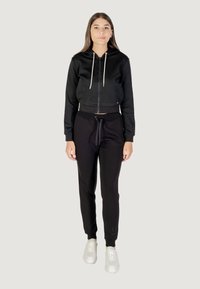 Felpa nera corta con zip frontale e cordini, abbinata a pantaloni jogger neri con vita elasticizzata e sneakers bianche.