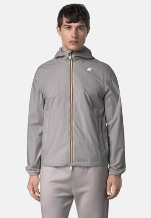 Giovane uomo con giacca grigia chiaro con cappuccio e cerniera, polsini elastici e pantaloni abbinati, in piedi contro uno sfondo semplice.