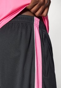 Μαύρες αθλητικές βερμούδες με υφή, διαθέτουν ροζ πλαϊνές λ ωρίδες με λευκή επένδυση. Ένας τσέπης με φερμουάρ είναι ορατός στη δεξιά πλευρά.