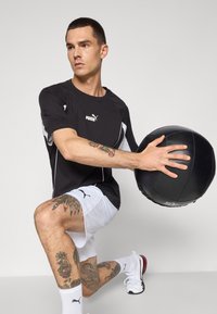 Černé sportovní tričko s bílými akcenty; model v bílých šortkách drží černou medicinbal. Sportovní obuv a viditelné tetování na nohách.