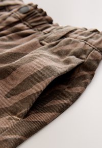 Tissu à motif camouflage marron avec une finition légèrement texturée ; comprend une ceinture élastique et un détail de poche latérale.
