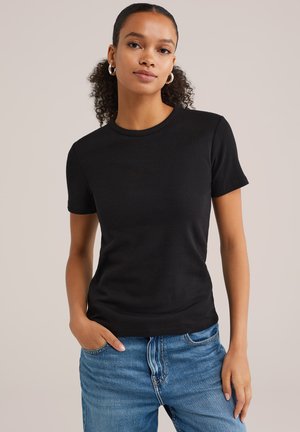 T-shirt nera aderente realizzata in tessuto liscio, scollatura rotonda, maniche corte, abbinata a jeans di denim blu chiaro, mani nelle tasche.