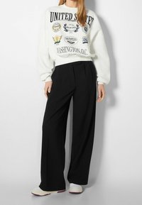 Biała bluza z napisem "STANY ZJEDNOCZONE" i grafiką; zestawiona z czarnymi spodniami o szerokich nogawkach i białymi sneakersami z fioletowymi podeszwami.