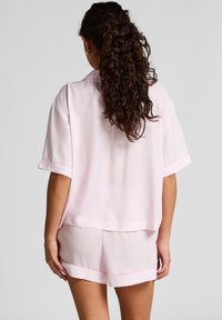 Vrouw met krullend haar, gekleed in een lichtroze T-shirt met korte mouwen en bijpassende shorts, staande met haar gezicht naar weg tegen een effen achtergrond.