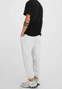 Bílé tepláky se bočními kapsami, v kombinaci s černým tričkem a šedo-bílými teniskami, pohled zezadu. Jednoduchý design, uvolněný střih.