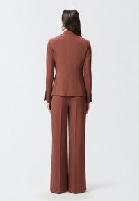 Smeđi odijelo po mjeri s prilagođenim sakoom i širokim hlačama. Glatka tkanina, zatvaranje na jedan gumb i čiste linije.