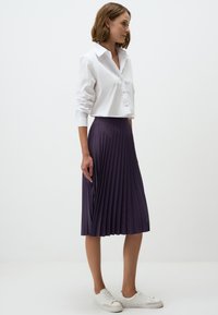 Camicia bianca con colletto abbinata a una gonna plissettata viola fino al ginocchio e scarpe da ginnastica bianche. Tessuto dalla consistenza liscia e design a strati.