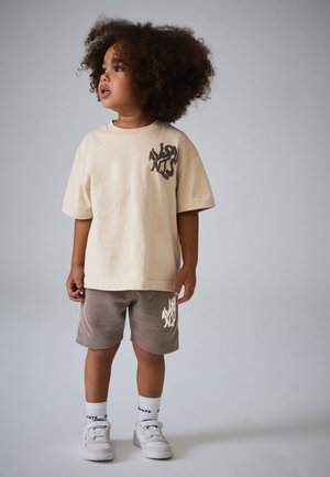 Young child with curly hair wearing beige graphic t-shirt, brown shorts, white socks, and sneakers, standing against a plain background.