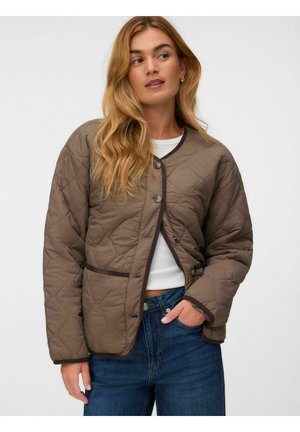 Kvinde med langt blond hår iført en brun quiltet jakke over en hvid top og blå jeans, der kigger til siden med den ene hånd i lommen.