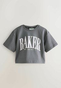 Tmavě šedé zkrácené tričko s krátkým rukávem s velkým bílým nápisem "BAKER" a menším nápisem "BY TED BAKER" na přední straně, položené na světlém povrchu.