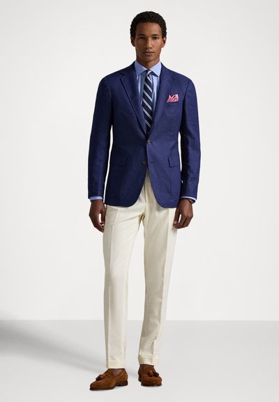 Vyras stovi apsirengęs tamsiai mėlynu švarku, šviesiai mėlyna marškinėliais, dryžuota kaklaraiščiu, kreminėmis kelnėmis, rudos spalvos šlepetėmis su kutais ir rožine kišenine nosine.