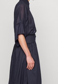 Henkilö, jolla on päällään tumma, löysä, puolielämytsistä materiaalia oleva paita, jonka hihat ylettyvät kyynärpäihin, ja yhteensopiva hame, kuvattuna sivusta yksinkertaista taustaa vasten.