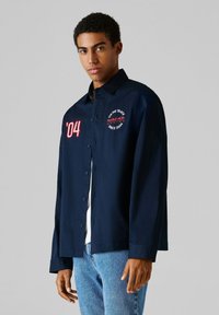 Νεαρός άνδρας που φοράει σκούρο μπλε πουκάμισο με κουμπιά, με τις επιγραφές '04' και Red Bull Racing, συνδυασμένο με ανοιχτόχρωμα μπλε τζιν, στέκεται μπροστά σε απλό γκρι φόντο.