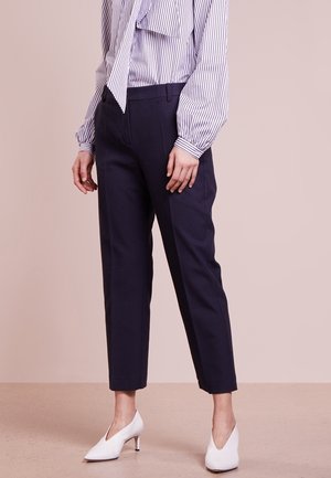 Kobieta ubrana w granatowe, dopasowane spodnie do kostek, białe szpilki oraz białą bluzkę z pionowymi granatowymi paskami i dużą kokardą przy szyi.