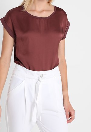 Femme portant une blouse en satin marron à manches courtes rentrée dans un pantalon blanc taille haute avec une ceinture nouée et des poches.