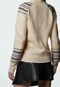 Sweater beige avec des manches à motifs marron, bleu et rose ; col côtelé ; porté sur une jupe en cuir noir ; texture douce.