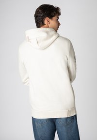 Helle cremefarbene Kapuzenjacke mit Kängurutasche, geripptem Saum und Bündchen. Verfügt über ein Logo in zartem Rosa auf der Kapuze und Schriftzüge an den Ärmeln.