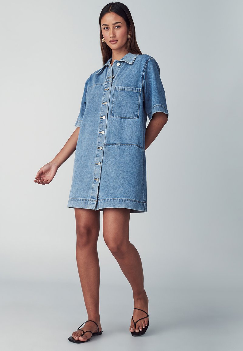 Denim jurk in lichtblauw, korte mouwen, sluiting met knopen aan de voorkant, enkele borstzak en een losse pasvorm, die tot halverwege het bovenbeen reikt.