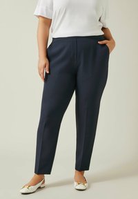 Marineblauwe op maat gemaakte broek met een gladde textuur, voorzien van zijzakken en een rechte pijp. Gepaard met witte flats met gouden accenten.