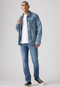 Lyseblå denimjakke med to brystlommer, båret over en hvit t-skjorte, kombinert med lyseblå jeans og svarte joggesko på en grå bakgrunn.