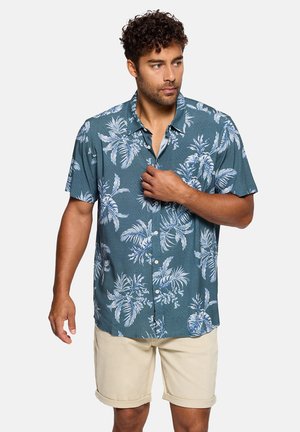 Hombre con camisa azul de manga corta con estampado blanco de hojas tropicales y pantalones cortos beige remangados, mirando hacia su derecha.