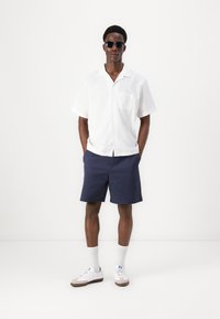 Hvit kortermet skjorte med lomme, marineblå shorts og hvite sneakers med grå detaljer, accessorert med svarte solbriller.