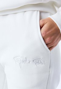Pantalones de chándal blancos hechos de una tela suave, con una cinturilla acanalada y bolsillos laterales, con el texto bordado en blanco "Project x Paris" en el muslo.