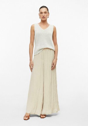 Femme debout de face, portant un débardeur en maille gris clair, un pantalon beige à jambes larges, des boucles d'oreilles créoles et des sandales brunes sur fond uni.