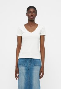 Γυναίκα με σκουρόχρωμο δέρμα και κοντά μαλλιά, που φοράει λευκή μπλούζα με V-λαιμόκοψη και κοντά μανίκια, καθώς και ψηλόμεσα μπλε τζιν, μπροστά σε απλό φόντο.