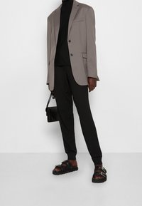 Person som bär en taupe blazer, svart turtleneck, svarta byxor, svarta sandaletter med spänne och håller en liten svart handväska.
