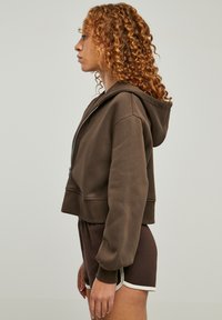 Brun cropped hettegenser laget av mykt stoff, med hette og glidelås. Kombinert med matchende brune shorts med hvit kant. Generelt avslappet passform.